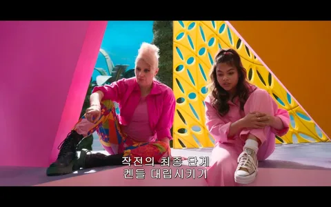 ```
In this vibrant video frame, two individuals are seated in a colorful, geometric environment. The person on the left is wearing a bright pink jacket and multicolored, patterned pants, paired with black combat boots. Their hair is styled in a distinctive, tall mohawk. They appear relaxed, with one arm resting on their knee. To the right, another person is dressed in a pink outfit, including a long-sleeved top and matching pants, accompanied by white sneakers with a visible brand logo. They have long, wavy hair tied back and are hugging their knees. The background is equally colorful, featuring pink and yellow geometric structures with blue accents, creating a playful and dynamic atmosphere. The mood seems casual as if they are in a friendly discussion. The subtitle in Korean reads, "작전의 최종 단계 켄들 대립시키기," which translates to "Final phase of the operation: Incite the Kens against each other."