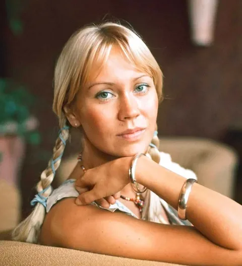Agneta Åse "Agnetha" Fältskog is depicted in this indoor portrait. She is seated in a relaxed posture, with her arm resting on a piece of furniture and her hand gently touching her chin. Her expression is calm and introspective, with a hint of a smile. Agnetha has blonde hair styled in braids, and she is wearing a sleeveless top which complements her casual yet polished look. Silver bracelets adorn her wrist, adding a touch of elegance to her appearance. The background is softly blurred, suggesting a cozy indoor setting with warm, muted tones. The lighting is soft and natural, highlighting her features and creating a serene atmosphere. This image captures a moment of quiet reflection, emphasizing Agnetha's graceful and poised demeanor.