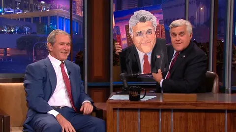 President George W. Bush is seated on the left side of the image, smiling in conversation. He is wearing a blue suit with a white shirt and a red tie, projecting a formal yet relaxed demeanor. To his right, Jay Leno is holding up a painting of himself. Leno is dressed in a dark suit with a white shirt and a dark tie, matching the formal setting of the talk show. The painting depicts Leno with an exaggerated smile, capturing the humorous tone of the setting. They are seated on a set that resembles a typical late-night talk show, with a desk in front of Leno and a cityscape backdrop visible through a large window behind them. The lighting is warm, suggesting an evening or nighttime broadcast. A black mug with "The Tonight Show" inscribed on it sits on the desk, reinforcing the setting as a television studio. The mood is light and jovial, fitting for a talk show segment featuring a former president.