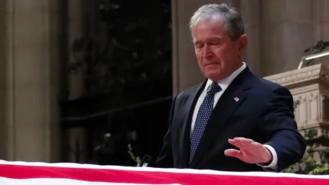 President George W. Bush is depicted in this image standing solemnly at a ceremony. He is in an indoor setting, likely a church or cathedral, given the ornate architecture visible in the background. The lighting is subdued, befitting a somber occasion. President Bush is wearing a dark suit with a white shirt and a patterned tie, appropriate attire for a formal event. He appears to be in a moment of reflection or mourning, as his expression conveys sadness and contemplation. In the foreground, there is a flag-draped casket, indicating this is part of a funeral service. The flag is prominently displayed, and its patriotic colors stand out against the more muted tones of the surroundings. The image captures a poignant moment, emphasizing the personal and national significance of the event.