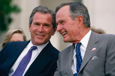 1) The image captures President George W. Bush and H.W. in a warm, candid moment outdoors. They are both dressed in formal attire, with George W. Bush wearing a dark suit, a white shirt, and a tie adorned with a pattern of small red and white elements. H.W. is dressed in a light gray suit with a white shirt and a blue tie featuring small dots. Both have short, neatly groomed hair. H.W. is wearing a pin on his lapel that resembles a flag, adding a touch of patriotism to his attire. The two are seated close together, engaged in a cheerful conversation, with smiles on their faces suggesting camaraderie and mutual affection. The background is softly blurred, indicative of a pleasant outdoor setting, possibly during a public event or gathering. Their relaxed posture and genuine expressions convey a sense of ease and shared history.
2) On-screen text block:
- Title: "..."