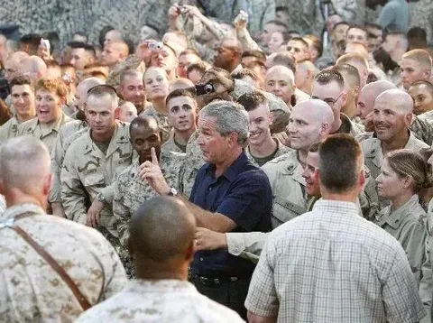 President George W. Bush is depicted in the center of a large gathering of military personnel. The scene is energetic, with many soldiers in uniform surrounding him. The soldiers appear attentive and some are smiling, capturing a moment of interaction and camaraderie. President Bush, dressed in a dark button-up shirt and light-colored pants, is gesturing with his right hand, possibly addressing the group or responding to someone in the crowd.
The atmosphere is bright, suggesting indoor lighting, and the setting feels informal yet organized. The soldiers are in various states of relaxed attention, with some holding cameras or phones to capture the moment. The background is filled with more soldiers, emphasizing the size of the gathering. The image conveys a sense of unity and morale, typical of a presidential visit to military troops. No specific text or branding is visible, focusing the attention on the interaction between President Bush and the soldiers.