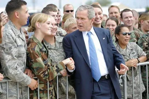 President George W. Bush is seen engaging with a group of military personnel. The setting is outdoors, likely on a military base or similar environment. President Bush is wearing a dark suit with a light blue tie, and he appears to be shaking hands and interacting warmly with the service members who are dressed in various military uniforms, including camouflage and digital patterns.
The mood is lively and positive, with several people smiling and appearing eager to meet the President. A barrier separates President Bush from the crowd, suggesting this is a formal visit or event. The lighting is natural, indicating it is daytime, and the background shows more people gathered, likely to see the President. The scene captures a moment of connection and mutual respect, typical of a presidential visit to military personnel.