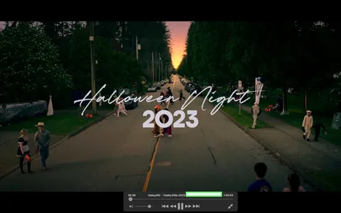 The image is a video frame capturing a festive Halloween evening in 2023. The scene takes place on a neighborhood street lined with trees and parked cars. In the foreground, people in various costumes are walking along the street. Some are dressed in whimsical or spooky outfits, creating a lively Halloween atmosphere. The lighting is dim, suggesting early evening as the sky glows with a hint of sunset in the background.
The image prominently features text that reads "Halloween Night 2023" in a stylized, handwritten font across the center of the frame, adding a thematic title to the scene. People are scattered across the street, some trick-or-treating, while others are engaged in conversations. The overall mood is festive and communal, typical of Halloween celebrations in suburban settings. The composition draws the eye toward the vanishing point down the street, emphasizing depth and the continuity of the celebration.