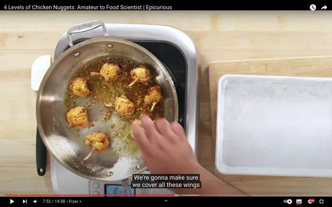 1) Rich description: The video frame is from a YouTube video titled "4 Levels of Chicken Nuggets: Amateur to Food Scientist | Epicurious," published by Epicurious. It captures a cooking demonstration in a kitchen setting. A stainless steel pan is on a stovetop, containing several golden-brown, fried chicken pieces, each with a skewer. The pan appears to have remnants of fried bits and oil, indicating the cooking process is well underway. A person’s hand, partially visible, is interacting with the food, likely turning or coating the chicken pieces. The background includes a wooden countertop with a cutting board and an empty metal tray on the side, suggesting preparation and organization for the cooking process. The lighting is bright, focusing on the food and hands-on action in the pan. The scene conveys a practical and instructional atmosphere, typical of cooking tutorial videos.
2) On-screen text block:
- Title: "4 Levels of Chicken Nuggets: Amateur to Food Scientist | Epicurious"
- Site / app: "YouTube"
- Captions / subtitles: "We're gonna make sure we cover all these wings"