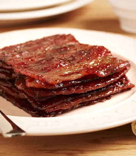 The image shows a close-up of a stack of bakkwa, a popular Asian snack. Bakkwa consists of thin, square slices of pork jerky that appear glossy and caramelized. The meat looks tender and slightly charred, indicative of its sweet and savory marinade. The bakkwa is arranged on a white plate, which contrasts with the rich, reddish-brown color of the meat. The lighting is warm, enhancing the appetizing appearance of the snack. The setting seems to be indoor, possibly on a dining table, with a hint of plates and a cup blurred in the background. The focus is primarily on the texture and glossiness of the bakkwa, making it the central element of the image.