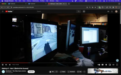 The image is a screenshot from a YouTube video titled "Making of - Halo 3 [Behind the Scenes]," uploaded by the channel "NeoGamer - The Video Game Archive." The scene is set indoors at a workstation with multiple computer screens. One of the screens prominently displays a scene from the video game "Halo 3," showing a gameplay environment with a character’s silhouette. Another screen appears to show graphic design software, indicated by a star-like shape. The third monitor displays a blank document or interface, suggesting work in progress.
The desk is cluttered with various items, including a keyboard, a Red Bull can, a red cup, papers, and some electronics. The lighting is dim, typical of an office space focused on computer work, possibly during a late-night development session. The atmosphere conveys a sense of concentration and creativity, commonly associated with behind-the-scenes looks at video game development.