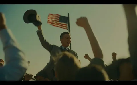 Cillian Murphy is depicted in this image from the film "Oppenheimer." He is surrounded by an enthusiastic crowd, raising their fists in celebration. Murphy, dressed in a suit and tie, holds a hat in one hand and is smiling broadly, conveying a sense of triumph or achievement. The scene is set outdoors, with an American flag waving prominently in the background, suggesting a patriotic or historical context. The lighting is warm and natural, giving the scene a vibrant and uplifting atmosphere. The composition focuses on Murphy, elevated above the crowd, highlighting his central role in the moment. The overall mood is jubilant, capturing a significant, victorious moment.