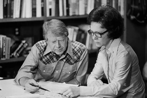 In this black-and-white image, Jimmy Carter is seated at a desk in an office setting, engaged in what appears to be a focused discussion. Next to him is a woman, both intently examining documents spread out in front of them. The background is filled with bookshelves, suggesting a well-stocked library or study environment. Jimmy Carter is dressed in a distinctive plaid shirt, which gives a casual yet professional appearance. The woman is wearing glasses and a light-colored blouse, her hair neatly styled. Both appear to be deeply concentrated on the task at hand, indicating a serious atmosphere. The lighting is even, highlighting the details of the room and the expressions on their faces. The presence of multiple papers and a pen in Carter's hand suggests they are involved in planning or reviewing detailed information.