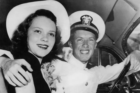 In this vintage black and white photograph, Jimmy Carter is seen sitting in a car, dressed in a naval officer's uniform, complete with a cap that features an insignia. He is smiling broadly, exuding a sense of happiness and pride. Beside him is a woman wearing a white hat and a floral dress, also smiling, suggesting a joyful and intimate moment between the two. The setting appears to be inside a car, possibly on a significant day such as a wedding or special event, given the attire and their expressions. The image captures a candid and timeless moment, showcasing the style and mood of the era. The lighting highlights their faces, creating a warm and nostalgic atmosphere typical of mid-20th century photographs. This image offers a glimpse into a personal and celebratory time in Jimmy Carter's life.