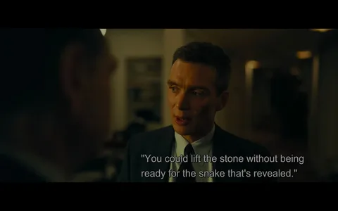 Cillian Murphy is featured in this scene from the film "Oppenheimer." The setting is indoors with subdued, warm lighting that creates a serious and intense atmosphere. Murphy is dressed in a formal dark suit and tie, suggesting a professional or high-stakes environment. His expression is focused and thoughtful, conveying deep contemplation or concern as he speaks. The background is softly blurred, indicating a room with minimal distractions, possibly an office or a meeting space. The scene captures a moment of dialogue, with the subtitle displaying, "You could lift the stone without being ready for the snake that's revealed." This metaphorical statement hints at underlying tensions or unexpected consequences in the storyline. The framing centers Murphy prominently, emphasizing the gravity of the conversation.