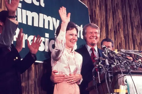 Jimmy Carter is seen in this image, standing behind a podium in a celebratory setting. He is smiling broadly, indicating a positive or triumphant moment, possibly during an election campaign or victory speech. Next to him is a woman, who is also smiling and waving to the crowd. Both are dressed in formal attire; Jimmy Carter is wearing a suit with a patterned tie, and the woman is in a light-colored blouse and skirt. The background features a partially visible sign with bold white text, "elect Jimmy" and "Presi," suggesting a political context. Multiple microphones are positioned in front of them, indicating a press event or public address. The scene is well-lit, contributing to the overall cheerful and lively atmosphere, and other individuals in the background are clapping, adding to the celebratory mood.