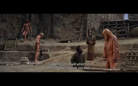 The image is a frame from the film "Planet of the Apes." It depicts a scene set in a rustic, outdoor environment, likely an archaeological dig site. Several characters are present, including humans dressed in primitive clothing and apes in more sophisticated attire. The apes are standing and interacting with the humans, suggesting a power dynamic or cultural exchange. The background features a rocky terrain with wooden structures like ladders and platforms. The lighting is natural, highlighting the earthy tones of the surroundings.
One ape, dressed in an orange and brown outfit, stands in the foreground conversing with another ape, who is lower and looking upward. A human male, also in primitive garb, is interacting with the environment to the left. The mood of the scene reflects a mix of curiosity and tension, as suggested by the subtitle: "suggests a culture which, in certain ways, equals our own." This indicates a thematic exploration of cultural parallels between the apes and humans.