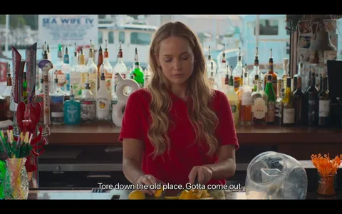 The image depicts a woman standing behind a bar counter, which is adorned with a variety of liquor bottles and bar equipment. She is wearing a red shirt and has long, wavy blonde hair, appearing focused and slightly pensive as she slices lemons on the counter. The background reveals a collection of bottles, including brands like Malibu and various other spirits, suggesting a well-stocked bar. To the left, a lobster decoration and colorful straws add a playful touch to the setting. The lighting is bright, indicating daytime, and provides a clear view of the objects and the woman's expression. Outside the window, a boat and water are visible, hinting at a coastal or waterfront location. Subtitles at the bottom read, "Tore down the old place. Gotta come out," suggesting a conversation or narrative moment. The overall mood is calm and somewhat introspective, typical of a scene where a character may be reflecting or engaged in routine work.