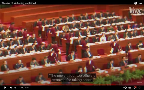 ```
This video frame from a Vox YouTube video titled "The rise of Xi Jinping, explained" depicts a formal governmental assembly, likely within a Chinese political context. The setting is a large, well-lit auditorium with rows of long wooden desks occupied by numerous attendees in suits, indicative of a serious political meeting or session. The scene captures a moment of significant activity with several attendants wearing maroon uniforms moving between the rows, distributing or collecting documents. The mood is formal and structured, reflecting the procedural nature of such gatherings.
In the foreground and background, individuals are seated in orderly rows, focused on paperwork or speaking with colleagues, suggesting engagement with official proceedings. The color palette is dominated by the rich reds and browns of the seating and desks, complemented by the dark suits of the participants, creating a cohesive and serious visual tone. The subtitles highlight a moment of political significance, referencing the removal of top officials for bribery, adding a layer of narrative tension to the scene. The presence of plants in the front suggests an attempt to soften the otherwise stern environment, typical of formal governmental settings.