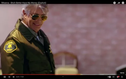 The image is a frame from the music video for Rihanna's song "Bitch Better Have My Money (Explicit)," available on YouTube. The frame shows a man with grey hair wearing sunglasses and a sheriff's uniform. The uniform has a patch that reads "COUNTY SHERIFF," suggesting his role in the scene. The background is slightly blurred, featuring a brick wall and some indistinct furniture, indicating an indoor setting. The lighting is warm, adding to the atmosphere of the video. The man appears to be smiling, which contributes to a somewhat casual or confident demeanor. The video is published by Vevo on Rihanna's channel. The hint indicates a connection to Julia Roberts, suggesting the man is her brother, though his identity is not explicitly confirmed in the frame.