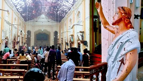 The image shows an indoor scene within a church that appears to have suffered significant damage. The left side of the image reveals a large group of people, likely congregants, standing and moving about amidst the chaos. The atmosphere is tense, as evidenced by the scattered debris across the floor and the distressed expressions and postures of the individuals present. The wooden pews are disordered, and the ceiling shows signs of damage, with exposed beams and broken sections.
On the right side of the image, a close-up view of a statue, likely representing a religious figure, is visible. The statue has been marked with what appears to be black stains or damage, adding to the somber mood. The figure is draped in a white robe and has one arm raised, possibly in a gesture of blessing or greeting. The scene is marked by a strong contrast between the serene expression of the statue and the surrounding disarray.
The lighting is dim, possibly due to the damage, lending a somber and serious tone to the overall image. The photograph captures a moment of aftermath, focusing on both the human response and the symbolic damage to a place of worship.