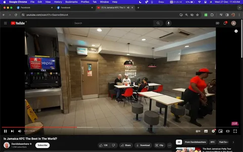 In this YouTube video screenshot titled "Is Jamaica KFC The Best In The World?" from the channel Davidsbeenhere, the scene takes place inside a KFC restaurant. The interior is modern, with tiled floors and wooden wall panels. On the left, there is a drink dispenser with a Pepsi logo. A group of four people sit at a table in the center-right, engaged in conversation while enjoying their meal. The group includes a person in a red shirt and hat, likely a staff member, who is standing and appears to be attending to the guests or cleaning. The lighting is bright and evenly distributed, creating a welcoming atmosphere. A sign on the wall reads "CHICKEN HAND-BREADED COOKED FRESH DAILY," emphasizing the restaurant's focus on fresh food. Red chairs and simple tables contribute to the casual dining setting.