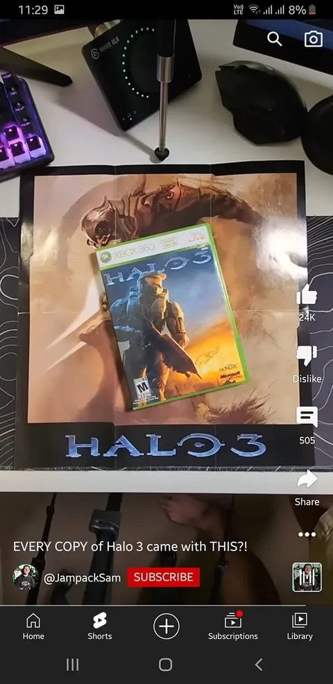 The image is a screenshot from a YouTube video titled "EVERY COPY of Halo 3 came with THIS?!" by the channel @JampackSam. The scene is set indoors on a desktop, featuring an Xbox 360 game case of "Halo 3" prominently displayed on top of a poster. The poster is mostly visible and depicts an armored character, likely from the game, with the "Halo 3" title clearly printed at the bottom. Surrounding the game and poster are various items typical of a gaming setup, including a colorful backlit keyboard, a microphone, a pair of headphones, and a device labeled "WAVE XLR." The lighting is bright, highlighting the gaming theme. The video seems to explore whether all copies of "Halo 3" included a specific item, possibly the poster shown. The layout and placement of objects suggest an emphasis on gaming culture and nostalgia for the Xbox 360 era.