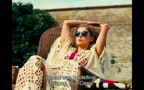 The scene shows a woman lounging outdoors, seated comfortably on a wicker chair. She is wearing sunglasses and a cream-colored crochet cover-up over a bright pink outfit, suggesting a summery and relaxed atmosphere. Her arms are stretched behind her head as she leans back, exuding a laid-back demeanor. The background consists of a stone wall and some lush greenery, indicating a garden or backyard setting. The lighting is bright and warm, typical of a sunny afternoon.
The subtitles reveal she is reminiscing about her modeling days, referencing cultural elements like Britpop, and bands like Blur and Oasis, which adds a nostalgic tone to the scene. This information hints at a discussion or reflection on past experiences in the fashion industry during the era when those bands were prominent.