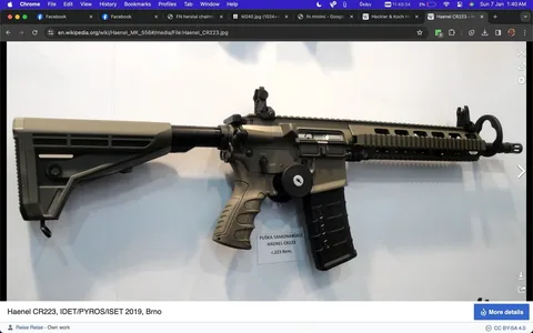 The image depicts a Haenel CR223 rifle displayed at IDET/PYROS/ISET 2019 in Brno. The rifle is mounted against a plain background, showcasing its design and features. It has a tactical appearance with a matte black finish and a collapsible stock. The rifle has a rail system on the upper receiver, allowing for the attachment of various accessories. The magazine is inserted, and the safety selector is visible near the grip. A tag hanging from the trigger guard provides information about the model and caliber: "PUŠKA SAMONABÍJECÍ HAENEL CR223 r .223 Rem." The image has a clean and professional look, typical of trade show displays, highlighting the technical and aesthetic aspects of the firearm.