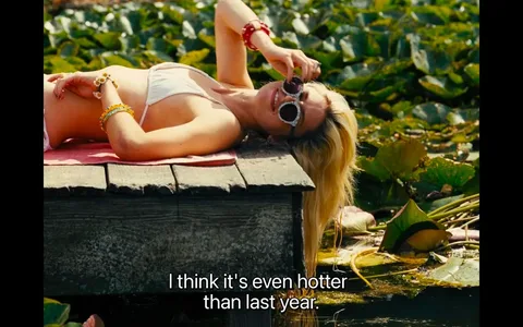 The image captures a relaxed summer scene with a person lying on a wooden dock beside a body of water filled with lily pads. The individual is wearing a white bikini and sunglasses, and they have long, blonde hair cascading over the edge of the dock. Their posture is laid back, and they appear to be enjoying the sunny weather, suggesting a warm, leisurely setting. The person is accessorized with colorful bracelets, adding a playful touch to their attire.
In the background, lush green lily pads dominate the scene, emphasizing the natural environment. The lighting is bright, indicative of a sunny day, casting warm tones over the entire image. The overall mood conveys a sense of tranquility and enjoyment of a hot summer day. The on-screen caption reads, "I think it's even hotter than last year," reflecting the character's thoughts on the temperature.