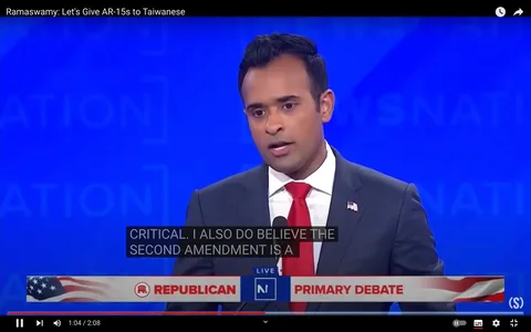 Vivek Ramaswarmy is featured in this video frame taken from a Republican Primary Debate. He is positioned in the foreground, speaking into a microphone. Ramaswarmy is wearing a dark suit with a white shirt and a red tie, with a small American flag pin on his lapel, indicating his participation in a formal political setting. The background is a solid blue with indistinct patterns, suggesting a large event or stage backdrop typical of televised debates.
The title of the video, "Ramaswamy: Let's Give AR-15s to Taiwanese," appears at the top, implying a controversial or significant statement made during the debate. The on-screen text includes a subtitle that reads, "CRITICAL. I ALSO DO BELIEVE THE SECOND AMENDMENT IS A," suggesting a discussion related to gun rights or foreign policy. Below Ramaswarmy, a banner with the text "REPUBLICAN PRIMARY DEBATE" is prominently displayed, adorned with an American flag motif. The overall mood of the scene is serious and focused, reflecting the formal and high-stakes nature of political debates.