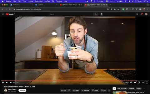 The image is a screenshot from a YouTube video titled "Julia Child's Crème Brûlée | Jamie & Julia" on the channel ANTI-CHEF. The setting is a modern kitchen with wooden countertops and dark cabinetry in the background. The scene shows a person leaning forward with both elbows on the countertop, holding a small ramekin in one hand and a spoon in the other, likely tasting the crème brûlée. The person is wearing a denim shirt and an apron, suggesting that they are the host or chef of the video. The lighting is warm, creating a cozy and inviting atmosphere. The YouTube interface is visible, showing a red progress bar at 6:25 of the 7:05 video. The number of subscribers, 376K, is displayed below the channel name. The video appears to focus on a dessert recipe, specifically crème brûlée, inspired by Julia Child.