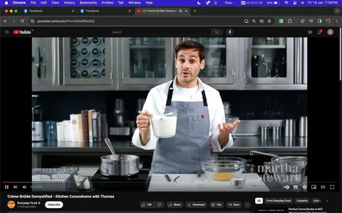 The screenshot captures a scene from a YouTube video titled "Crème Brûlée Demystified - Kitchen Conundrums with Thomas" on the Everyday Food channel. The setting is a modern kitchen, featuring stainless steel cabinets and various kitchen equipment. Thomas is standing behind a counter, wearing a white shirt and a gray apron, holding a measuring cup filled with cream. His expression seems informative and engaged, as he explains the process of making Crème Brûlée. In front of him, there are several bowls and a saucepan on a stove, suggesting he is in the midst of a cooking demonstration. The lighting is bright, focusing on Thomas and the ingredients. The video is published by "martha stewart," and is themed around cooking and baking, specifically focused on desserts. On-screen text includes "martha stewart," located at the bottom-right corner of the image.