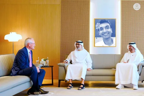 In this image, three men are seated in an elegantly designed room, engaged in a conversation. The decor features a modern aesthetic with warm wooden tones and a soft glowing lamp on the left. The man on the left wears a blue suit and a pink tie, sitting on a couch facing the two other men in traditional attire. The middle man, dressed in a white kandura and sandals, smiles as he converses with the man on the right, who is also in traditional attire and appears to be holding papers. A framed black-and-white painting of Lehan hangs prominently on the wall behind them, adding a personal touch to the setting. The atmosphere is formal yet relaxed, with elements like bottled water and a small flower arrangement on the table emphasizing the setting's hospitality. The lighting is bright and even, highlighting the clean and polished interior.