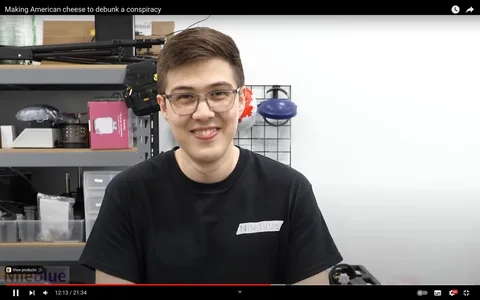 The image is a frame from a YouTube video titled "Making American cheese to debunk a conspiracy" on the channel NileBlue. It shows a person indoors, possibly in a workshop or lab setting. The individual is smiling, wearing glasses, and a black t-shirt with a homemade label reading "NileBlue." Behind them is a shelving unit with various items including boxes, tools, and safety gear, suggesting a workspace environment. The lighting is bright, typical of an indoor setting, which highlights the subject’s face. The overall mood is casual and engaging, aligning with the exploratory and educational theme of the video. No captions are visible on the screen.