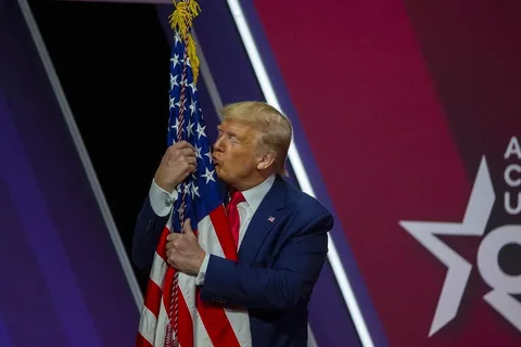1) Rich description: The image shows Trump at a public event, embracing and kissing an American flag. He is dressed in a dark blue suit with a white shirt and a red tie, projecting a formal and patriotic demeanor. The background features a stylized star design with the partial text "AC" and "USA," likely indicative of the event's theme or sponsor. The setting is indoors, with a stage and a decorative backdrop that uses bold colors such as purple and red, adding to the ceremonial atmosphere. The flag is held tightly by Trump, and his expression is one of affection and reverence. The lighting is bright and focused, illuminating Trump and the flag prominently, creating a focal point for the audience and the photograph. The composition centers on Trump's interaction with the flag, symbolizing a display of national pride.
2) On-screen text block:
- Title: "AC USA"
