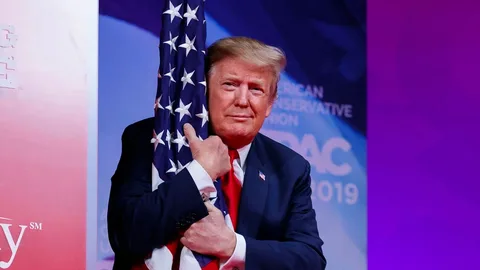 Donald Trump is depicted in this image, embracing an American flag on a stage. The setting appears to be a conference or event, indicated by the backdrop with the text "AMERICAN CONSERVATIVE" and "CPAC 2019," suggesting it's from the Conservative Political Action Conference. Trump is wearing a dark suit with a red tie, and he is standing with a composed expression, embodying a sense of patriotism. The lighting is bright, emphasizing the vivid colors of the flag and his attire. The stage background has a gradient of red and blue tones, contributing to the theme of the event. The atmosphere is formal yet personal, with Trump’s gesture signifying a connection to national symbols. The image captures a moment where the focus is on Trump and the flag, reflecting political themes and his public persona.