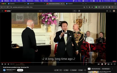 The image is a screenshot from a YouTube video titled "South Korean president sings ‘American Pie’," published by the Washington Post. The scene takes place indoors, likely at a formal event or gathering. In the foreground, a man with a microphone is dressed in a black tuxedo with a white shirt and bow tie, suggesting a formal occasion. Behind him stand several individuals in military uniforms, indicating a ceremonial or official atmosphere. A large floral arrangement and ornate decor are visible, adding to the formal setting.
In the left part of the image, another man, whose back is to the camera, seems to be listening to the singer. The setting is well-lit, with warm lighting reflecting off the white walls and elegant furnishings. A piano is partially visible, hinting at the musical nature of the event. The subtitle on screen reads, "A long, long time ago," a line from the song "American Pie," which the South Korean president is performing. The mood appears to be celebratory and engaging, as evidenced by the attention of the audience members.
- Title: "South Korean president sings ‘American Pie’"
- Channel / profile: "Washington Post"
- Site / app: "YouTube"
- Captions / subtitles: "A long, long time ago"