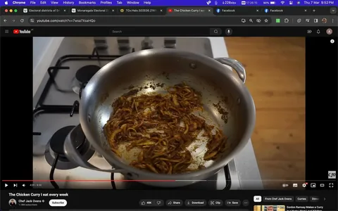 The image is a screenshot from a YouTube video titled "The Chicken Curry I eat every week" by Chef Jack Ovens. It shows a cooking scene in a kitchen setting. In the foreground, there is a large, stainless steel pan on a stovetop, with a mixture of sautéed onions and spices beginning to caramelize. The pan is positioned on a gas burner, suggesting active cooking. The stovetop is clean and modern, with black burner grates and knobs visible. The onions in the pan have a rich, golden-brown color, indicating they are being cooked for a flavorful base, likely for a curry dish. The lighting is bright, highlighting the texture and color of the ingredients in the pan. The kitchen surface around the stovetop is tidy, emphasizing a focus on the cooking process. At the bottom of the screenshot, YouTube interface elements are visible, including control buttons and the channel name "Chef Jack Ovens," which indicates the content creator's focus on cooking tutorials and recipes. The video timestamp is at 4:09 of a total 8:00 minutes, suggesting this is a mid-process step of the recipe.