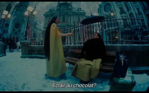 Emma Stone is featured in this scene from the film "Poor Things." The setting is an outdoor area with a snowy ground, giving it a wintry ambiance. The architecture in the background suggests a historical or fantastical setting, with grand buildings and intricate designs. Emma Stone is wearing a long, flowing yellow cape, and her dark hair is notably long. She is standing and appears to be offering something, possibly an éclair, to a man sitting on a bench. The man is wrapped in dark clothing, and a suitcase is placed beside him on the snow-covered ground. The lighting is soft, creating a serene yet mysterious atmosphere. The entire scene is stylized with a slightly surreal, almost dreamlike quality, typical of a fantasy period film. The subtitle reads “Éclair au chocolat?” indicating a light, possibly humorous moment.