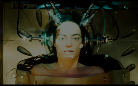 Emma Stone is depicted in a scene from the film "Poor Things," where she appears to be in a futuristic or scientific setting. She is lying down with her head encased in a transparent dome connected by several metal and rubber tubes, suggesting a theme of experimentation or medical intervention. The setting is dimly lit, with a focus on her face, highlighting her expressionless and slightly vacant stare. The lighting casts a cool, clinical glow, enhancing the sci-fi atmosphere. The background includes various metallic and mechanical components, emphasizing a laboratory-like environment. The mood is tense and surreal, with Emma Stone's character appearing vulnerable yet central to the scene's focus on technology and human experimentation.