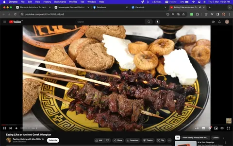 The screenshot is from a YouTube video titled "Eating Like an Ancient Greek Olympian" on the "Tasting History with Max Miller" channel. The image shows a plate filled with various foods that reflect ancient Greek cuisine, aiming to recreate the diet of an ancient Olympic athlete. The centerpiece of the plate is skewered meat, likely lamb or beef, grilled and seasoned with herbs. Accompanying the skewers are slices of rustic bread, a block of cheese, and a pile of dried figs, all set on a plate with a decorative Greek pattern. The scene is set indoors with bright lighting that highlights the textures and colors of the food. There is no visible branding or text aside from the YouTube interface, which shows options like "Share," "Download," and "Clip" prominently displayed.