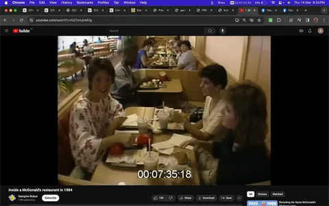 ```
The video frame captures a moment inside a McDonald's restaurant in 1984. Three people are seated at a table, enjoying their meal. The woman closest to the camera is wearing a white top with a red floral pattern and is engaged in a conversation. She appears cheerful and is gesturing slightly with her hands as if emphasizing a point. The two other women at the table are listening attentively; one is wearing a white top and the other a dark-colored outfit. The table is laden with typical fast-food items: a burger, fries in the iconic red McDonald's packaging, and drinks with straws.
The setting is warm and nostalgic, characteristic of 1980s interior design, evident in the wooden booth seating and simple decor. In the background, other patrons are visible, suggesting a busy lunchtime environment. Bright natural light filters in from the windows, creating a lively and inviting atmosphere. The timestamp "00:07:35:18" is visible on the screen, indicating the video’s runtime. The video is hosted on YouTube under the channel "Vampire Robot," which often revisits historical or vintage themes.