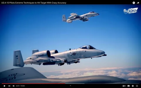 The image is a frame from a YouTube video titled "US A-10 Pilots Extreme Techniques to Hit Target With Crazy Accuracy." It features two A-10 aircraft in flight against a clear blue sky, highlighting their role in precision targeting. The aircraft are flying in close formation, demonstrating coordination and skill. The foreground shows one A-10 Warthog with distinct features such as its twin turbofan engines and a nose painted with the emblem of the U.S. Air Force. The tail of another aircraft is visible, marked with "MI" and "184," indicating its squadron or unit. The lighting is bright, with the aircraft casting subtle shadows, emphasizing their aerodynamic shapes. The video is published by the channel "fluctus," known for content related to aviation and military technology. The visual composition captures the elegance and power of these specialized warplanes, with a focus on their operational capabilities in a controlled airspace.