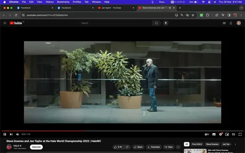 Steve Downes is featured in this screenshot from a YouTube video titled "Steve Downes and Jen Taylor at the Halo World Championship 2022 | HaloWC." The video is hosted on the HALO channel. The scene is set indoors with a modern, nighttime ambiance. Steve Downes is standing by a row of large potted plants, seemingly engaged in a phone conversation. He is dressed casually in a dark jacket, jeans, and boots, and is positioned to the right of the frame. The setting is spacious and well-lit, with a glass barrier and a muted color palette contributing to a serene atmosphere. The image conveys a moment of contemplation or communication, likely relevant to his involvement with the Halo series. The video is related to the "Halo 3" game, adding context to the interaction depicted.