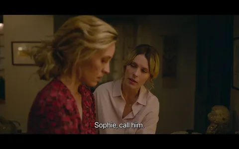 In this image, two women are engaged in a serious conversation inside a warmly lit room, suggesting an intimate or private setting. The woman on the left, possibly Sophie, wears a red patterned blouse and looks focused on something out of frame, her expression pensive. The woman beside her, dressed in a light-colored striped shirt, is speaking to her, appearing concerned and supportive. Her posture leans slightly towards Sophie, indicating empathy or encouragement. The lighting emphasizes the warm, cozy atmosphere of the scene, with soft shadows adding depth to their expressions. The subtitle, "Sophie, call him," suggests a moment of decision or emotional tension, indicating that Sophie's action is significant to the narrative. The background is blurred, focusing attention on the interaction between the two women.