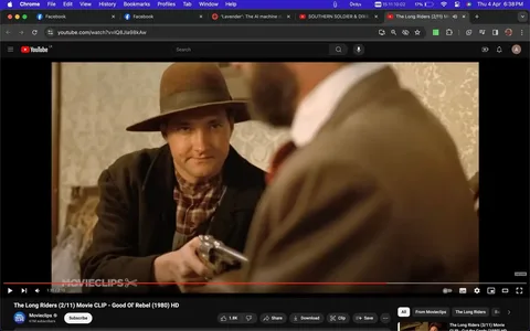 The screenshot is from a YouTube video titled "The Long Riders (2/11) Movie CLIP - Good Ol' Rebel (1980) HD," uploaded by the channel "Movieclips." The scene is from the film "The Long Riders." It captures a moment inside a room, where a man wearing a wide-brimmed hat and a plaid shirt is holding a revolver. His expression is slightly amused or confident, and he is seated at a table, engaging with another person whose back is visible in the foreground. The lighting is warm and natural, highlighting the period-appropriate attire and setting indicative of a Western film. The wallpaper in the background suggests a modest, possibly rustic environment. The composition draws focus to the man with the gun, emphasizing tension or a pivotal moment in the narrative. On-screen text includes the video title and channel information, along with subscription prompts and related video suggestions. The lack of visible captions suggests direct engagement with the visual and auditory elements of the clip.