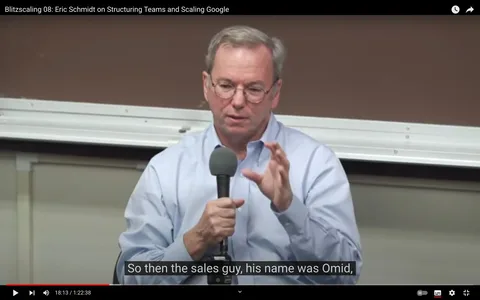 Eric Schmidt is seen speaking in a video frame from the YouTube channel titled "Blitzscaling 08: Eric Schmidt on Structuring Teams and Scaling Google." He is holding a microphone with both hands, using a hand gesture to emphasize his point. Eric Schmidt is wearing glasses and a light blue button-down shirt, seated in an indoor setting that looks like a lecture hall or conference room, visible from the whiteboard in the background. The lighting is bright and even, typical of an indoor setting designed for presentations or discussions. The subtitles at the bottom of the screen read, "So then the sales guy, his name was Omid," suggesting he is sharing an anecdote or insight related to sales within a corporate setting. The atmosphere is formal yet engaging, indicative of a professional talk or lecture.