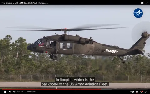 The image is a frame from a video titled "The Sikorsky UH-60M BLACK HAWK Helicopter" on YouTube. It depicts a Sikorsky UH-60M Black Hawk helicopter in flight, hovering above a tarmac or runway area, with greenery visible in the background. The helicopter is dark green and has the words "UNITED STATES ARMY" emblazoned along the side with the registration number "N600PV" prominently displayed. The word "EXPERIMENTAL" is also visible on the helicopter's side, indicating that this might be a test or prototype model. The rotors are spinning, suggesting the helicopter is either taking off or landing. The overall lighting is bright, indicating a daytime setting. The helicopter appears to be mid-motion, captured with slight motion blur on the rotating blades, providing a dynamic feel to the image. Subtitles at the bottom mention the helicopter's role as the "backbone of the US Army Aviation Fleet."