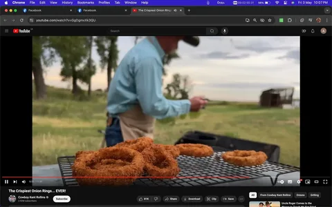 In this screenshot from a YouTube video titled "The Crispiest Onion Rings… EVER!", the scene captures a rustic outdoor setting. The focus is on a pile of crispy onion rings placed on a metal rack, suggesting they have just been freshly fried. The foreground is dominated by the golden-brown onion rings, emphasizing their crunchy texture. Slightly blurred in the background, a person—presumably Cowboy Kent Rollins—wears a light blue shirt, jeans, and a cowboy hat, embodying a quintessential Western look. He seems to be preparing or handling something out of focus, perhaps related to the cooking process. The surrounding environment includes grassy fields and distant trees, adding to the rustic and natural ambiance. The lighting is soft, with a slightly overcast sky visible, indicating an early morning or late afternoon setting.
- Title: "The Crispiest Onion Rings… EVER!"
- Channel / profile: "Cowboy Kent Rollins"
- Site / app: "YouTube"