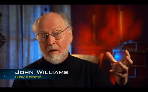 The image depicts John Williams, identified as a composer, during what appears to be an interview. He is seated indoors, in a warmly lit room with a wooden backdrop that suggests a study or library. His attire is casual, consisting of a black turtleneck sweater, and he is wearing glasses. John Williams has an expressive demeanor, with one hand raised as if he is explaining or emphasizing a point. The lighting is soft, creating a cozy atmosphere, and the background includes a large, partially visible globe, adding to the scholarly feel of the setting. The text identifying him as "John Williams" and "Composer" is prominently displayed at the bottom of the image, confirming his identity and role.