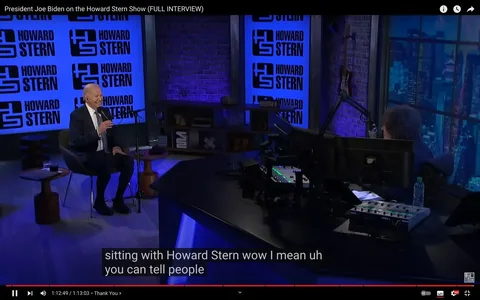 In this scene, President Joe Biden is featured as a guest on the Howard Stern Show. The setting is an indoor studio, characterized by its contemporary design with a blue and black color scheme. President Biden, seated on the left, is smiling and holding a microphone, engaged in conversation. He is wearing a dark suit with a white shirt and patterned tie. The background features multiple screens displaying the "HOWARD STERN" logo in bold, black lettering against a blue backdrop, creating a branded environment. On the right, Howard Stern is seated behind a large desk with multiple computer monitors and audio equipment, typical of a radio or talk show setting. The lighting is dim, focusing on the participants, enhancing the intimate interview atmosphere. The studio also includes shelving with various decorative items and a guitar hanging on the wall. The caption on the screen captures a moment from their conversation, further emphasizing the informal and conversational tone of the show.
- Title: "President Joe Biden on the Howard Stern Show (FULL INTERVIEW)"
- Site / app: "YouTube"
- Captions / subtitles: "sitting with Howard Stern wow I mean uh you can tell people"