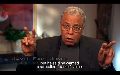 James Earl Jones is featured prominently in an interview setting. He is wearing glasses, a dark blazer, and a grey shirt, conveying a sense of professionalism and formality. In the image, he is using air quotes with both hands, suggesting emphasis on the phrase he is discussing. The background is softly lit and out of focus, featuring some decorations like a plant and a framed painting, contributing to a cozy atmosphere typical of an interview set.
The on-screen text identifies him as "JAMES EARL JONES," and he's credited as the "VOICE OF \"DAR," likely referring to a notable character he has voiced. His expression is serious yet thoughtful, indicative of a reflective conversation. The subtitle reads, "but he said he wanted a so-called 'darker' voice," hinting at a discussion about voice acting and character portrayal. The lighting is warm, highlighting James Earl Jones's face and adding to the intimate feel of the interview.