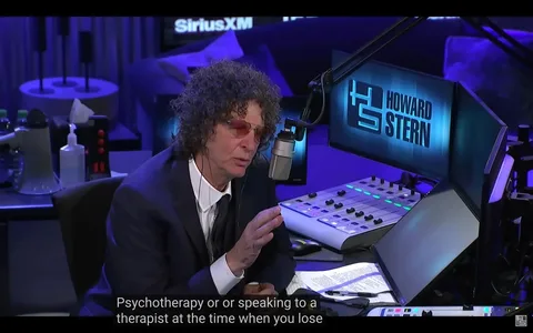 Howard Stern is pictured in a radio studio, likely hosting his show on SiriusXM. The setting is equipped with professional broadcasting equipment, including a large microphone positioned in front of him, a soundboard, and multiple computer monitors. The studio has a dimly lit, bluish ambiance that gives a modern and technological feel. Howard Stern is wearing a suit with a tie, and his expression is focused and engaged, suggesting a serious discussion. His curly hair and distinctive style are characteristic of his persona. To his left, there are bottles and a pump bottle of sanitizer on the table. The background features a prominent display with "Howard Stern" and "SiriusXM" logos. The subtitles at the bottom mention "Psychotherapy or or speaking to a therapist at the time when you lose," indicating a conversation likely centered around mental health or personal development.